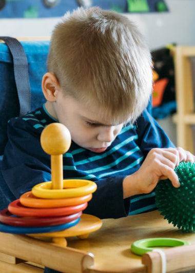A young child sits in a supportive chair, holding a green sensory ball. Colorful toys are on the tray, with more toys on shelves behind. Image
