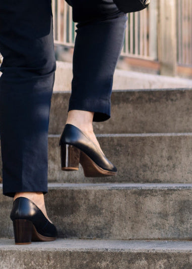 A person wearing black heeled shoes and dark pants is ascending a set of concrete stairs. A metal railing runs alongside the stairs. Image
