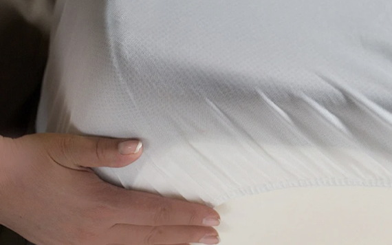 A close-up of a person stretching a white fitted sheet over a mattress corner, smoothing it for a snug fit. The sheet is soft and smooth.