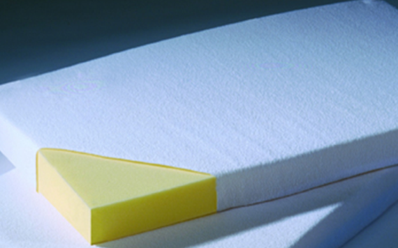 A close-up of a mattress corner shows yellow foam inside, with a white textured fabric cover against a plain background.