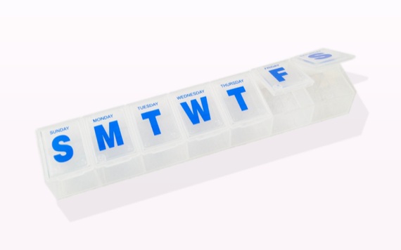 A white pill organizer with blue letters showing days Sunday to Saturday, each day has its own openable compartment.