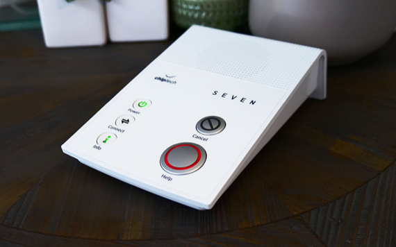 A white medical alert device with buttons for power, connect, cancel, and help. It has green/red indicators and SEVEN label on wood surface.