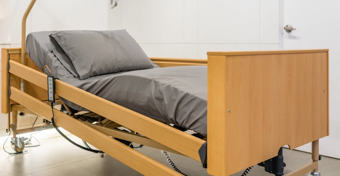 A hospital bed with a wooden frame and gray sheets sits near a white wall in a minimalist room with a closed door and nightstand.