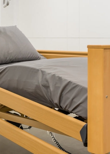 A hospital bed with a wooden frame and gray sheets sits near a white wall in a minimalist room with a closed door and nightstand. Image