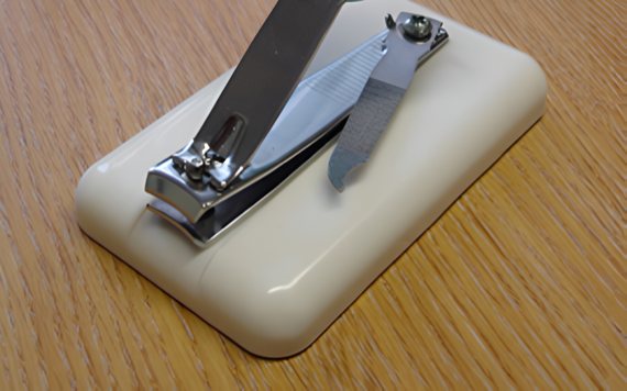 A metal nail clipper with a built-in nail file rests on a curved, off-white plastic holder on a wooden surface.
