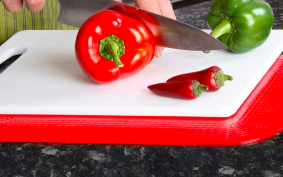 A person slices a red bell pepper on a bordered white board with small chili peppers and a green bell pepper, using a large knife.
