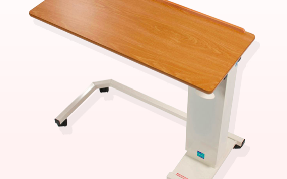 A height-adjustable overbed table with a light brown wooden surface and a white metal base, designed for hospitals or home care.