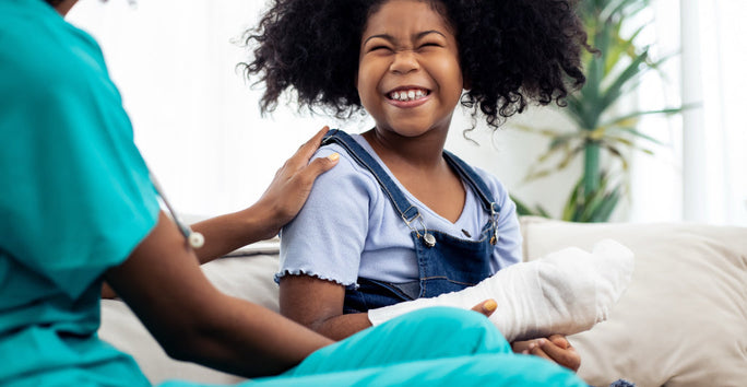 Smiling young girl with a bandaged arm sitting on a sofa while a caregiver offers reassurance and support.