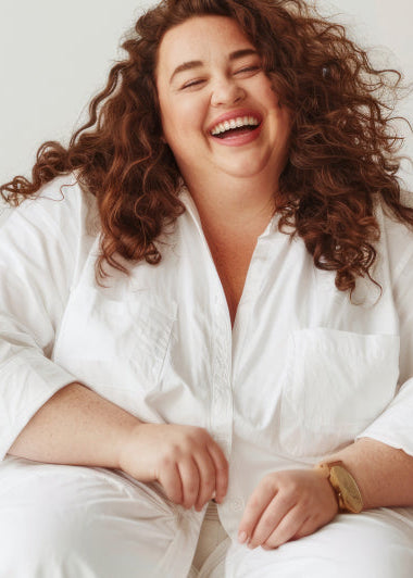 A person is happily sitting on a wooden chair, wearing a white outfit, in front of a light-colored wall. Image