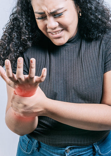 A woman with a distressed expression holds her right wrist, highlighted by a red glow to show pain, wearing a dark top and jeans. Image