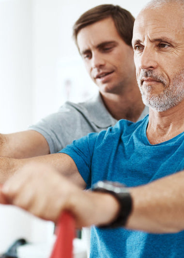 An older man uses a resistance band for exercise, assisted by another man in a well-lit gym or therapy room. Both concentrate on the task. Image