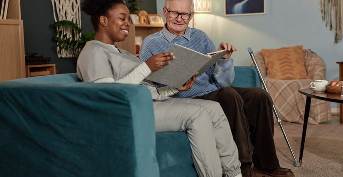 A caregiver in a grey uniform sits with an elderly man on a blue couch, smiling at a photo album together in a cozy, warmly lit room.