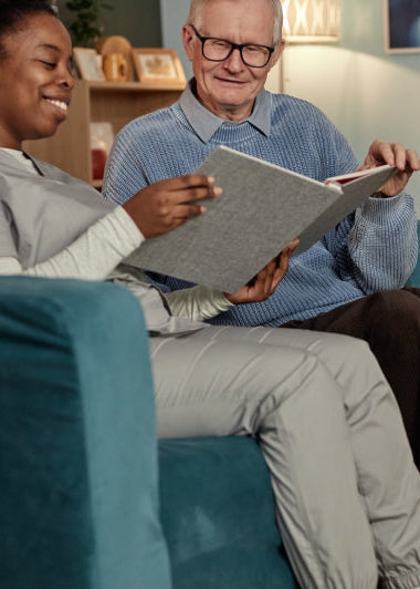 A caregiver in a grey uniform sits with an elderly man on a blue couch, smiling at a photo album together in a cozy, warmly lit room. Image