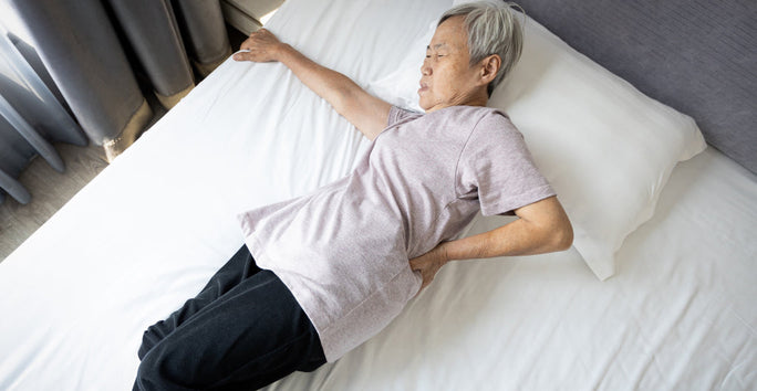 An elderly person lies on a bed, wearing a light purple shirt and dark pants, resting with one hand on their lower back.