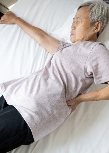 An elderly person lies on a bed, wearing a light purple shirt and dark pants, resting with one hand on their lower back. Image