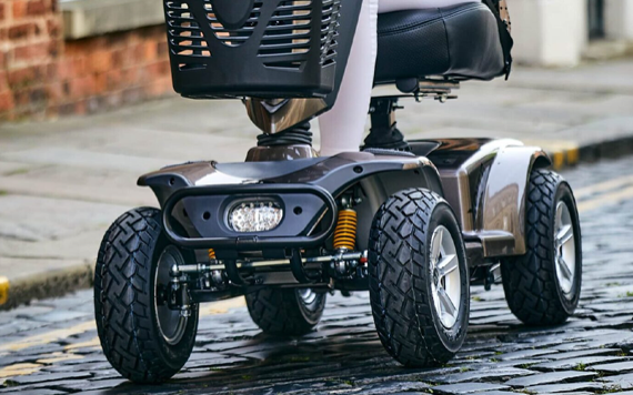 Close-up of a mobility scooter on cobblestones with large tires; a person in white pants sits on it, visible from the knees down.