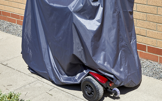 A mobility scooter is partly hidden by a blue tarp on a paved path next to a brick building. The red rear wheel area is visible.