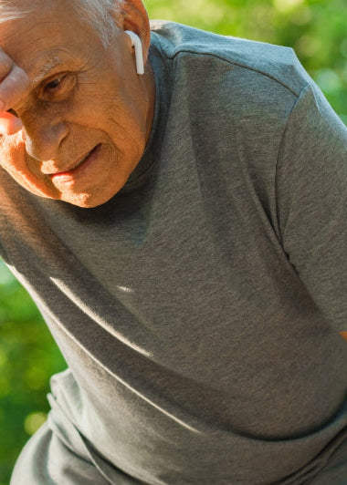 An elderly man stands outside, wearing earbuds and a gray T-shirt. He wipes his forehead, looking tired, against a background of green trees. Image
