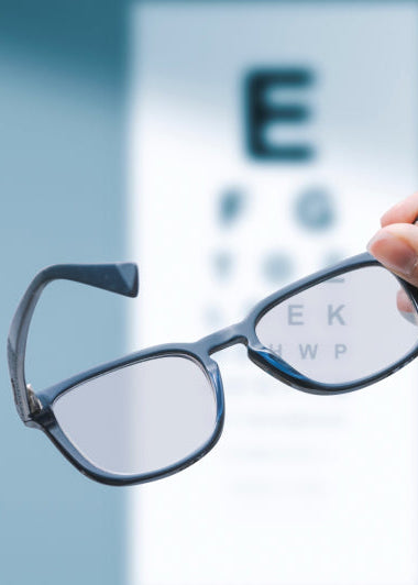A hand holding eyeglasses in front of a blurred eye chart with letters, against a blue background. Image