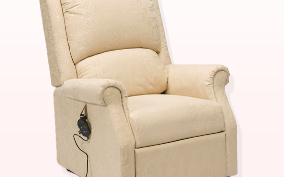A tan recliner with a high back and cushioned arms sits against a light pink backdrop. A remote on a black cord is attached to the side.