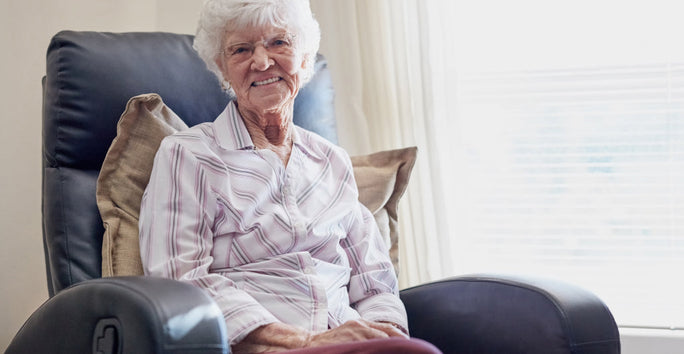 An older woman wearing glasses sits in a cozy chair, smiling. She wears a striped shirt; a bright room with a window is behind her.