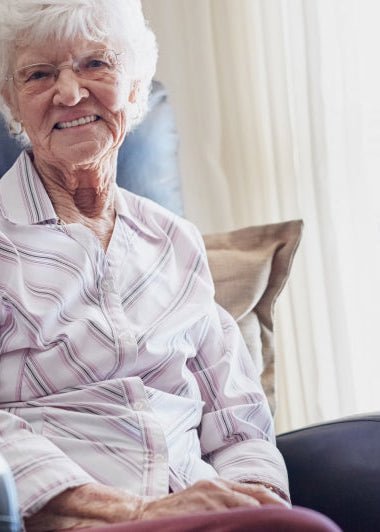 An older woman wearing glasses sits in a cozy chair, smiling. She wears a striped shirt; a bright room with a window is behind her. Image