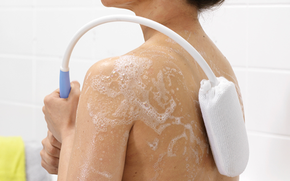 A person uses a long-handled scrub brush to wash their back, covered in soap suds. The white bathroom tiles form the background.