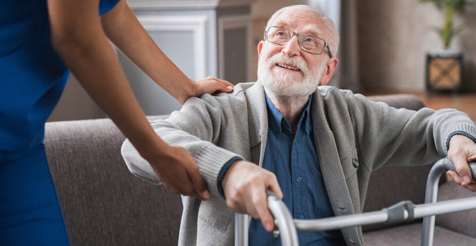 An older man smiles while using a walker, helped by a person in blue scrubs indoors with a couch and plant nearby.