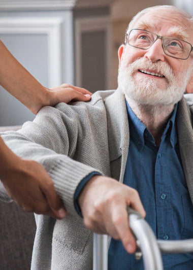 An older man smiles while using a walker, helped by a person in blue scrubs indoors with a couch and plant nearby. Image