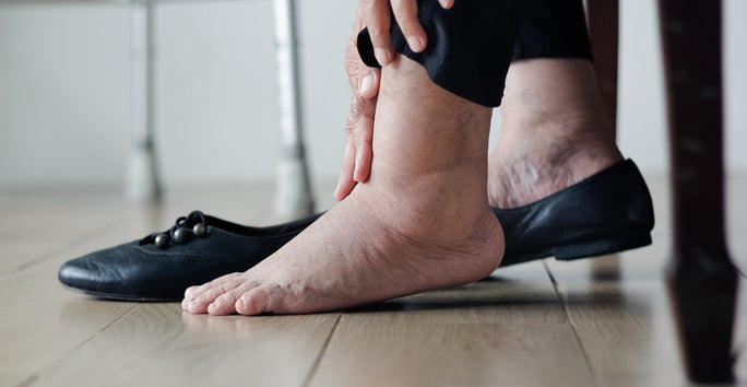 A person is sitting, holding their swollen ankle. One shoe is off, foot bare on a wooden floor, indicating discomfort.