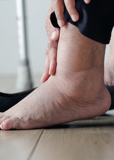 A person is sitting, holding their swollen ankle. One shoe is off, foot bare on a wooden floor, indicating discomfort. Image
