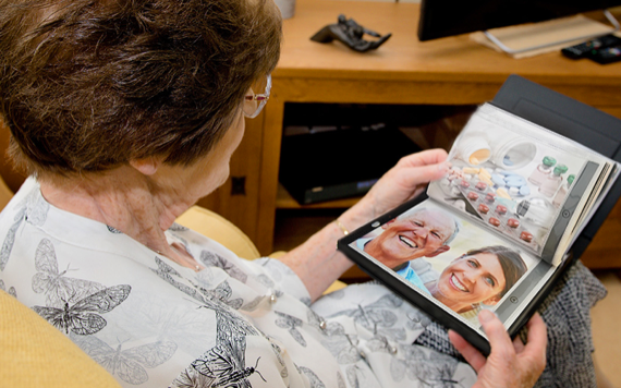 An older woman in a butterfly-patterned blouse looks at a photo album with family pictures next to images of pills on the page.