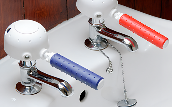 A white sink with shiny chrome taps and colored handles, set against brown wooden paneling. A plug chain is attached to one tap.