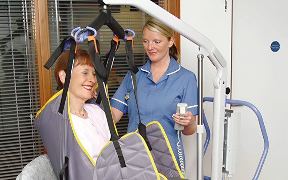 A caregiver helps a seated woman using a hoist in a medical room with blinds and equipment. The mood is supportive and caring. Image