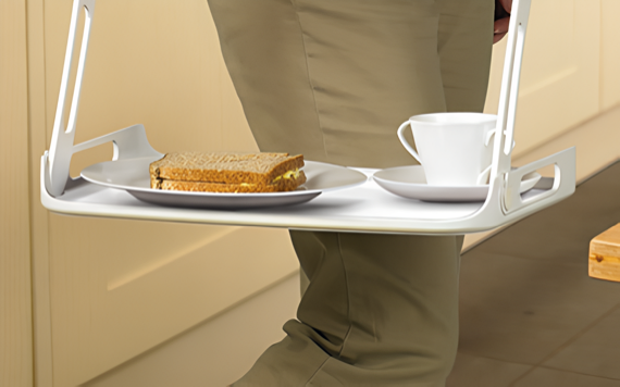 A person stands on a tiled floor, holding a white tray with a sandwich and cup. Light cabinets are in the background.