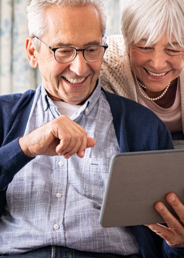 An older couple smiles at a tablet, man in glasses on couch, woman leans over. They look happy in a cozy living room. Image