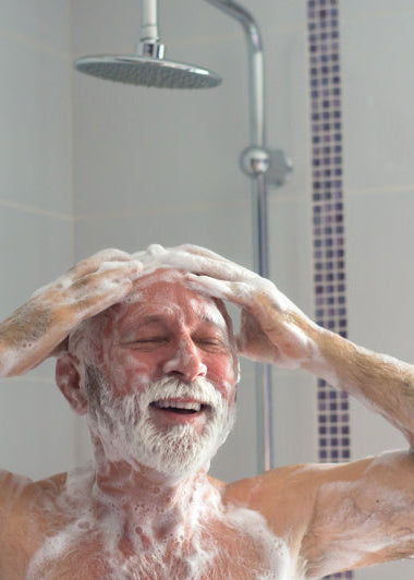 An older man smiles under a shower, covered in soap, washing with both hands. Light tiles and modern showerhead visible. Image