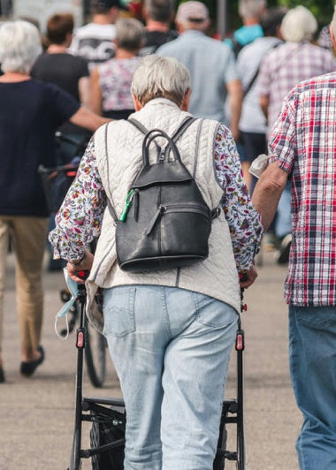 An elderly couple walks on a busy street; the woman uses a walker, the man wears a plaid shirt, and colorful flowers line their path. Image