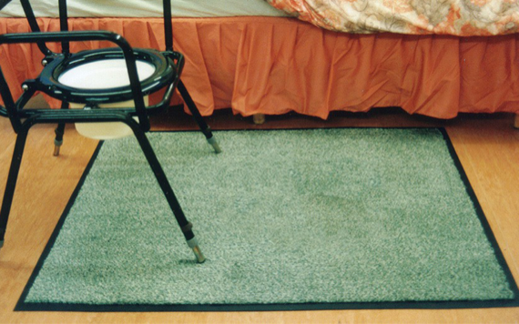 A metal commode chair with a white seat sits on a green rug on a wooden floor next to a bed with a red skirt.