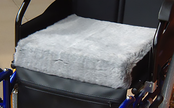 Close-up of a wheelchair with a soft, thick light gray fur cushion on the seat. The frame is black with some blue elements.