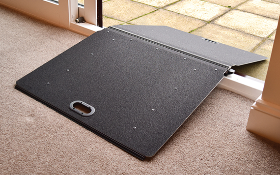 A dark gray folding ramp connects a carpeted indoor space to a tiled outdoor area, featuring a handle and textured surface for traction.