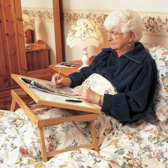 Adjustable wooden bed tray with legs in use by elderly woman lying in floral bedding, crossword open on tray, side view.