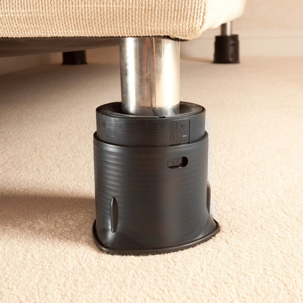 Bed leg with Langham SureGrip in a black furniture raiser lifting the bed on a beige carpet, close-up view.