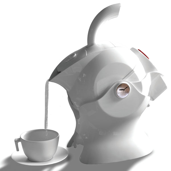 White Uccello Designs kettle with ergonomic handle pouring into a teacup on white surface, side view.