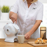 White Uccello Kettle with ergonomic handle, woman pouring tea next to scones and coffee, in use view.