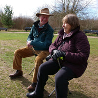 Two older adults sitting on red Medequip Ta-Da Chairs outdoors, trees behind them, chairs in use, front view.