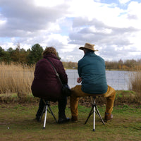 Two people using Medequip Ta-Da Chairs with red seats by a lake, sitting in tall grass, outdoor use view.