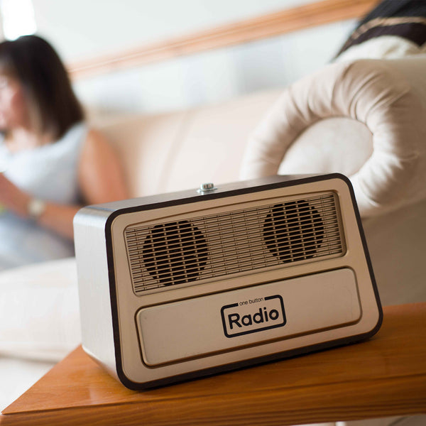 Ravencourt One Button Radio on a wooden table, woman sitting on beige sofa in soft focus background, front view.