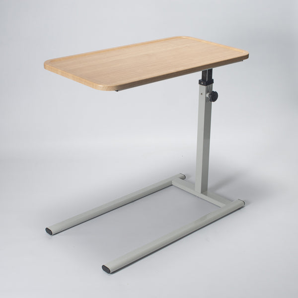 Height adjustable overbed table with light wood top and grey base, shown beside a bed, angled view.