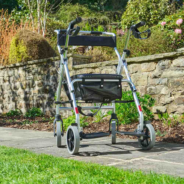 NRS 2 in 1 Rollator and Transit Chair with padded seat and bag on a garden path, stone wall and plants in background, angled side view.